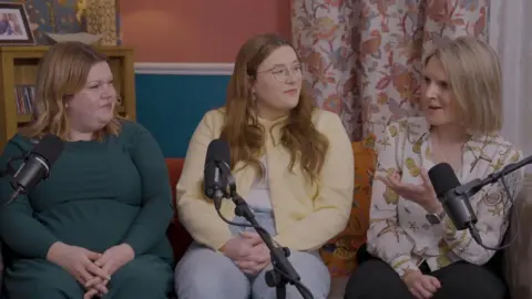 Lime Pictures Three women - Natasha, Jess and Marie-Claire - are sitting on a couch with microphones in front of them. The room is decorated with brightly-coloured floral wallpaper. 