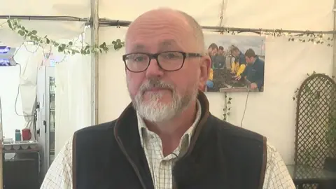 Chris Wright stands with glasses on, a short beard and a shirt with a jacket over the top of it.