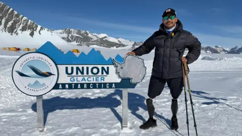 Hari Budha Magar standing at a sign for Union Glacier in Antarctica, near to Mount Vinson. Hari is wearing a cap, sunglasses, puffer jacket and shorts. He is holding two walking sticks. 