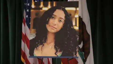 Reuters A photo of Celeste Rivas Hernandez is displayed on an easel in front of the US and California flags onstage at a media conference announcing charges against US singer David Anthony Burke, who is known professionally as D4vd.
