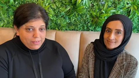 Farzana Jamidar and Raheela Hussain sit together in the cafe. Farzana has her long dark hair tied back and is wearing a black hoodie, Raheela is wearing a brown tweed jacket and black headscarf.