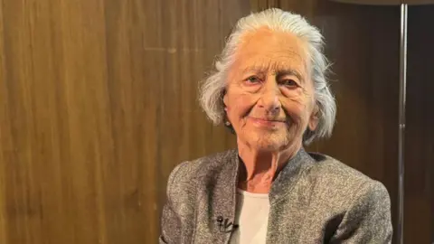 BBC/Mark Denten Susan Collingwood-Cameron, an elderly woman with short grey hair, her head and shoulders visible. She is smiling with her mouth closed. She is wearing a grey smart jacket on top of a white t-shirt. There is a wooden wall behind her.