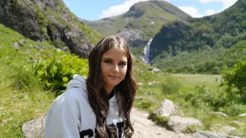 Ellie, who has long, brown hair and is wearing a grey hoodie, is sitting in a glen surrounded by fields and mountains