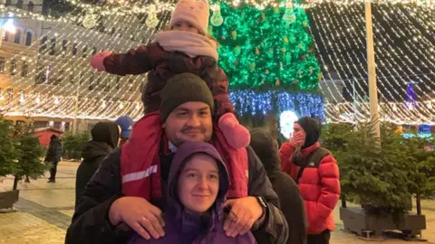 Iryna Mezenina A woman, man and little girl pose in front of a Christmas tree in a square decorated for Christmas