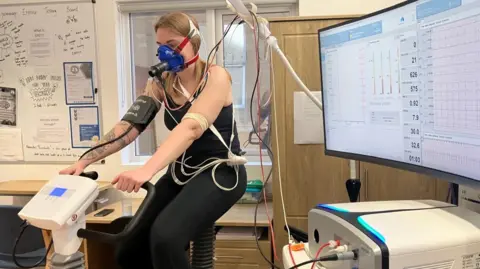 Sophie Pierce sits on a static bike in a hospital office room. She is wearing a black vest and black leggings and has a mask over her face which is hooked up to multiple cables. A monitor on her righthand side shows her vital statistics.