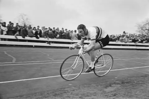 Getty Images A black and white image of a man on a racing bike on a circular track. He is wearing tight cycling shorts and top and a helmet and is leaning forward in a racing position. Standing crowds several people deep watch from behind a barrier at the outside of the track.