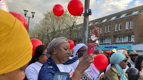 Shariqua Ahmed/BBC Crowds of people standing with red balloons in their hands.