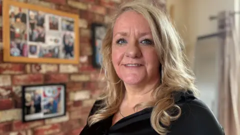 Simon Thake/BBC A woman with shoulder length blonde hair stands in front of a brick wall in a lounge with framed pictures on