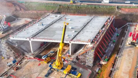 HS2 Ltd A large box-like structure with beams or girders on top sits on a building site next to a road. A large yellow crane stands next to it.