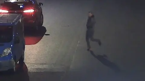 COPFS Hunter, in grey clothing, is walking away to the left of frame. His face is seen though it is grainy thanks to the CCTV image.