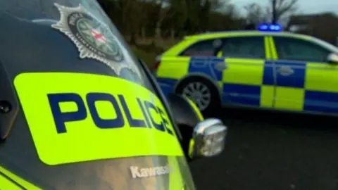 A police motorbike is seen in the foreground, with the word police in yellow type, written across the shield and a crest of the police service of Northern Ireland also visible. In the background is a yellow and blue PSNI car