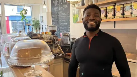 A man with dark hair and beard, wearing a black top, stands behind a coffee shop counter. 