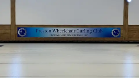 Blue sign saying "Preston Wheelchair Curling Club - Improve, Compete and Have Fun!" is on the wall above the ice.