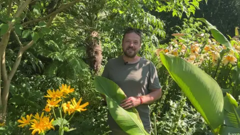BBC/NATHAN TURVEY Jack Ogg from York Gate Garden standing by his banana plant, he is wearing a green t-shirt and has brown hair and a beard, there are trees, flowers and shrubs around him.