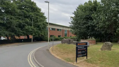 A curved road with double yellow lines leads towards a partially obscured building surrounded by trees. On the right, a grassy verge features large rocks and a black sign reading “RENSHAW” in orange, with “Renishaw plc” and contact details in white beneath.