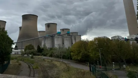 A general view of the decommissioned Cottam power station from the view of a railway bridge showing tracks in the foreground and a number of large cooling towers in the background. The sky is grey and cloudy. 