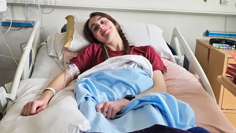 Bethany Handley Bethany lies in a hospital bed. Her long hair is plaited and she wears a dark red top. She has blankets covering her, a cuff around her upper right arm with tubes coming from it and a hospital identity bracelet on her wrist. There is a stand with medical monitors and wires to the left of the bed in the picture. To the right of the picture are a chest of drawers and a movable hospital table with a number of books on it. 