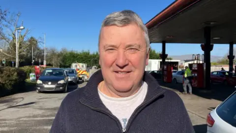 Martin White is smiling at the camera. He is wearing a navy knitted zip up and a grey t-shirt. He has short grey hair and is standing in a petrol station forecourt. 