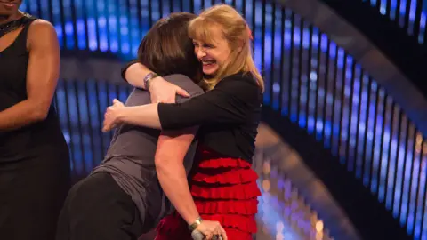 A picture of Liz Kenworthy hugging London bombings survivor Martine Wright on stage at the BBC's Sports Personality of the Year. Ms Kenworthy is wearing a red ruffled dress and black cardigan. Ms Wright is wearing dark trousers and a dark grey T-shirt. Ms Kenworthy is smiling broadly.