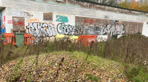 The Elmbridge Eagles site on Oaken Lane, Claygate. The club building is covered in graffitti and a number of pebbles can be seen in the image. The area is overgrown with weeds.