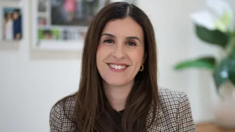 Macquarie University A woman with long dark hair and hoop earrings, wearing a check blazer, smiles at the camera.