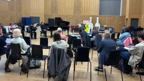A woman reads aloud from a sheet of paper as a small group of people on seats watch on.