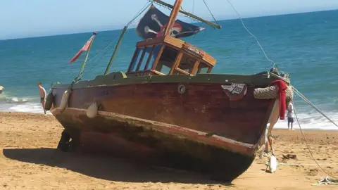 West Bay Discovery Centre The wooden vessel tilted at 45 degrees on the beach. It has a central wheelhouse and a mast bearing a pirate skull and crossbones flag.