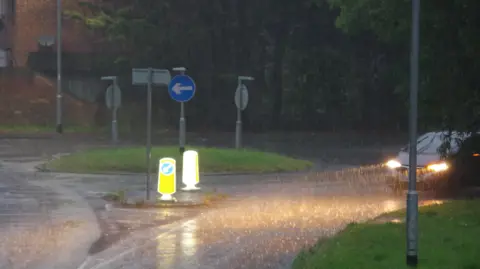 TUESDAY - A rainy day in Lower Earley shows a car with headlights on in downpour as it drives around a roundabout