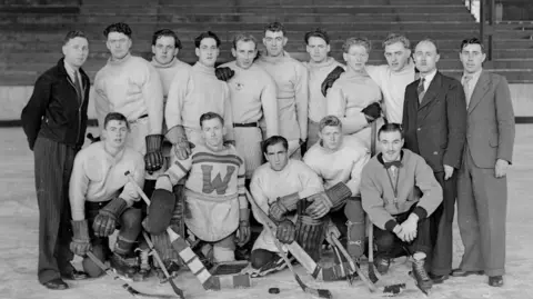 A black and white group photograph of Durham Wasps ice hockey team at Durham Ice Rink, October 1947. Russ Proudfoot is standing the back, third person from left to right. He has black wavy hair and is wearing a white jumper.