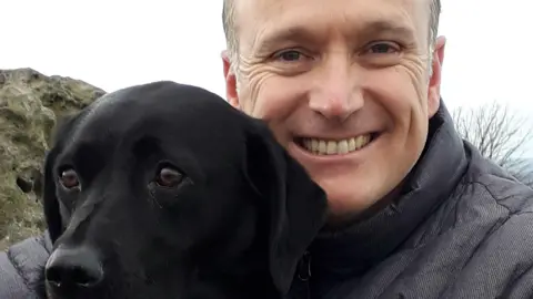 Supplied A man grinning while hugging a black dog, who is looking elsewhere