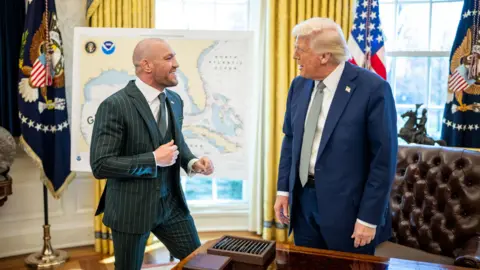 The White House Conor McGregor in a green suit in the Oval Office with blonde-haired Donald Trump, who is wearing a navy suit and a green tie.