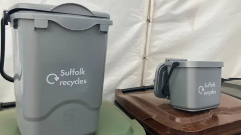 Vikki Irwin/BBC A large grey food caddie and smaller grey food bin sit on top of largest waste bins. The words Suffolk Recycles are printed on their fronts. They each have a black handle. 