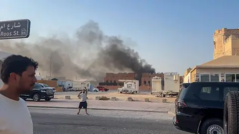 AFP via Getty Images A screenshot from a video shows smoke billowing from the roof of a building in the distance in Doha. The video is taken from a parallel road to where the smoke is appearing from, on which a man appears to be gesturing with his hands and another is located standing side-on nearer the camera. 