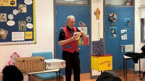 David Oakley the magician stands in front of a group of people. You can see the back of parents and children's heads. He is holding two large cards. One has a rabbit on it and the other has a black magician's hat on it. Next to him is  yellow and red box that is labelled "a box of tricks"