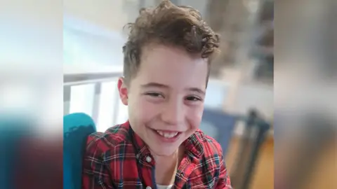 Nihat family Smiling young boy. He is wearing a chequered shirt and is looking at the camera. He has brown hair.