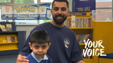 A father with dark hair and a beard is wearing a blue top. He has his right hand on his son in a school and they are both looking at the camera.