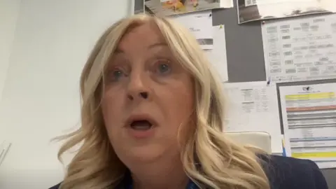 A middle aged woman with wavy, blonde hair speaks into a camera. She is wearing a blue blazer and is sitting in an office in front of a notice board.