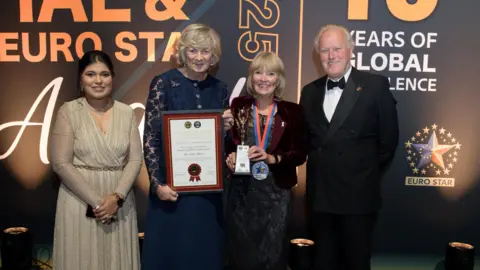Vox PR Women in silver dress, women in blue dress holding a certificate, a woman with a medal and a man in a tuxedo. They are standing in an indoor setting at an awards ceremony.