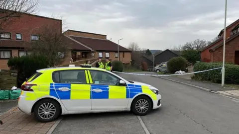 Three uniformed police officers stood on a residential street with a police tape cordon behind them and a police car parked in front of them. Behind the cordon are several brown houses, each with a driveway and green bushes in front of them.