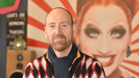 Wayne Saunders smiles at the camera. He has a ginger beard and short, cropped hair. He is wearing a navy scarf and black-and-red, diamond-patterned cardigan.