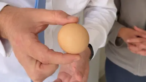 Simon Balch A man holds a spherical egg between his thumb and index finger. Another person stands nearby with their hands in front of their stomach and fingers interlinked. 