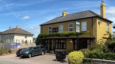 A large rectangular building is painted mustard yellow and sits in the side of a road with two cars parked outside. There is greeery on the front of the building and several windows over two floors.