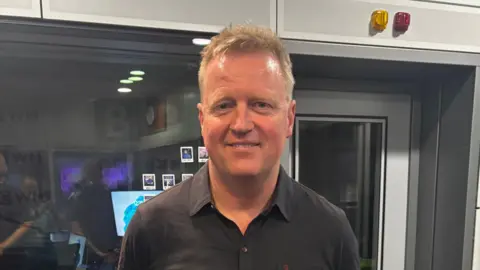 A photograph of a man wearing a black shirt looking directly itno the camera. There is a glass window and door behind him.
