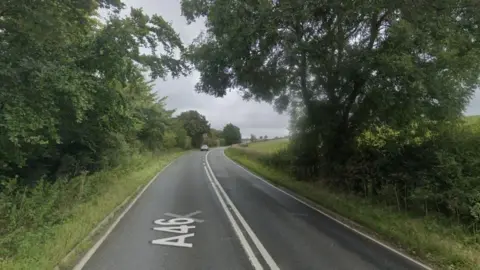 A Google Maps screengrab showing an empty A-road surrounded by trees, hedges and fields. 