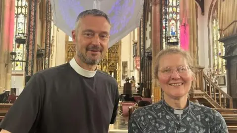 Maddy Jennings/BBC The Reverend Canon Edward Carter is on the left of the image. He has a lack top on and white collar around his neck. He has short grey hair and a short grey beard. On the right of the image, the Reverend Dr Haworth is looking into the camera, smiling. She has grey hair, tied back, and is wearing glasses. Her shirt is grey and patterned, and she has a white collar around her neck.