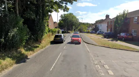 Google Cars driving along Hellesdon Road. Terrace houses line one side of the road and trees the other