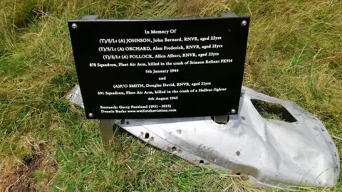 Jonny McNee A memorial for the Petty Officer, on a black plaque with white writing, it sits above a small silver piece of wreckage. The sign states the names of those killed in the crash and their ages.