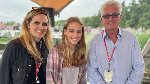 Jon Wright/BBC Sarah Lilley, Anna and Melvin Benn standing on the Waterfront Stage. They are all smiling at the camera.