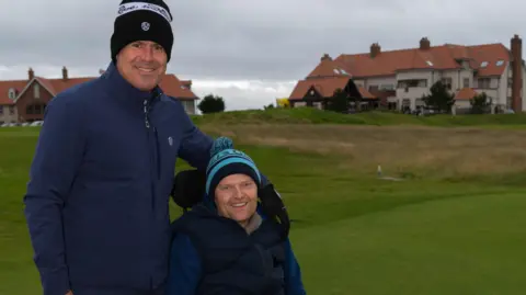 Kenny Logan stands with his hand on Kenny Scott's shoulders. Scott is sitting in a wheelchair. They are both wearing woolen caps and are on the golf course.