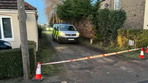 A police van and a car are parked in a dirt track road next to a bungalow and another house. There are traffic cones and a red and white cordon taping off the road